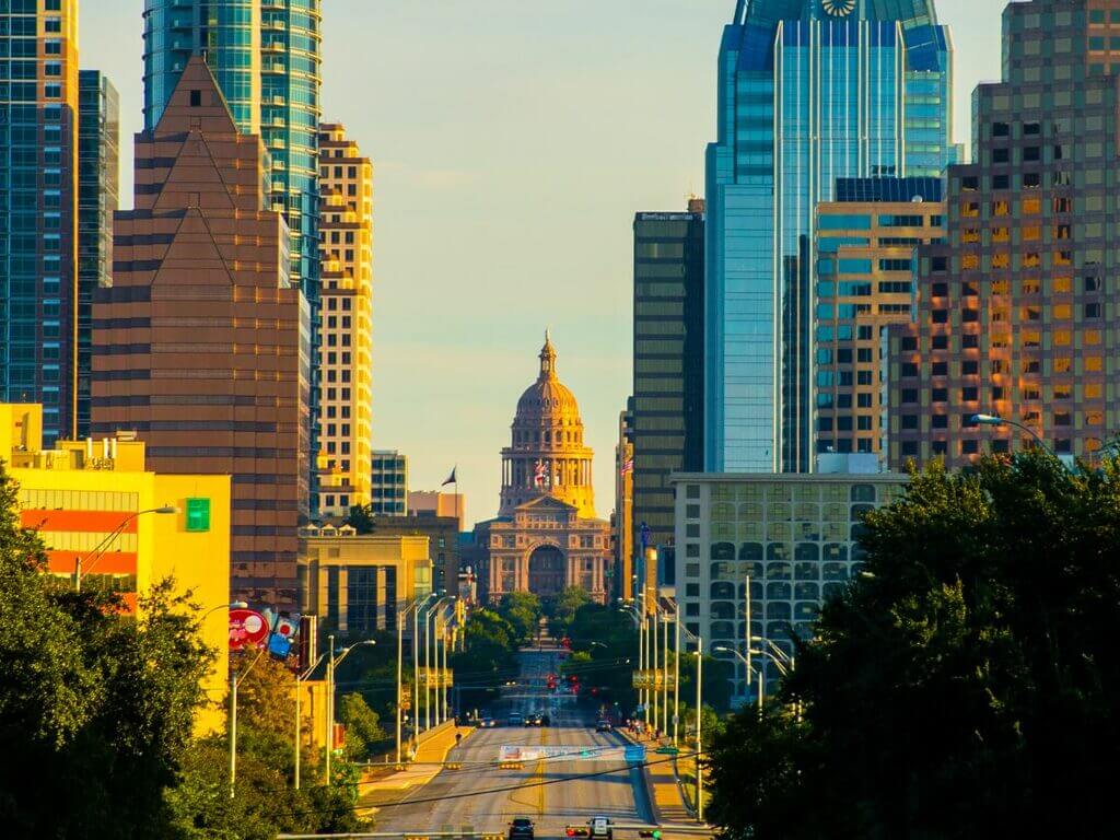 The Texas State Capitol building stands prominently at the end of a wide avenue, framed by modern high-rise buildings on both sides in downtown Austin.